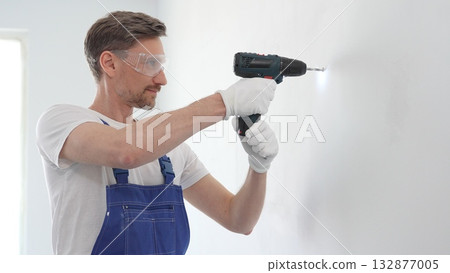 Professional handyman wearing blue overall, safety glasses and gloves, is using cordless drill to make hole in white wall during renovation or installation work 132877005