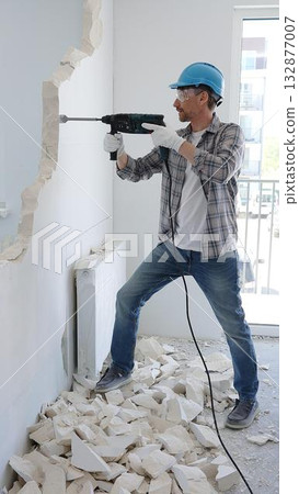 Man construction worker demolishing a wall with a hammer drill in protective gear, creating construction debris on the surrounding floor surface. Renovation concept 132877007