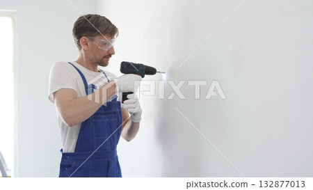 Middle aged male skilled worker drilling hole in white wall, wearing protective gear during home improvement project with cordless power tool Middle aged male skilled worker drilling hole in white wall, wearing protective gear during home improvement project with cordless power tool 132877013