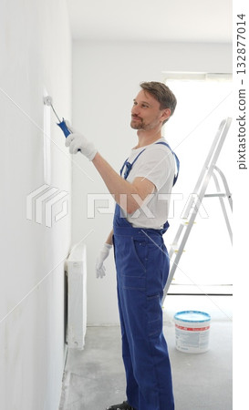 Middle aged man wearing protective white gloves and blue work overalls, applying paint with roller on pristine interior wall during home renovation project, vertical view Middle aged man wearing protective white gloves and blue work overalls, applying paint with roller on pristine interior wall during home renovation project, vertical view 132877014