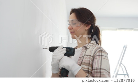 Woman wearing protective gear drilling hole in white wall during home improvement project, focused on precise home renovation work Woman wearing protective gear drilling hole in white wall during home improvement project, focused on precise home renovation work 132877016
