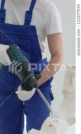 Close up of unknown male construction worker wearing blue overalls is demolishing white wall with rotary hammer drill, generating dust, close up horizontal view. Renovation concept Close up of unknown male construction worker wearing blue overalls is demolishing white wall with rotary hammer drill, generating dust, close up horizontal view. Renovation concept 132877019