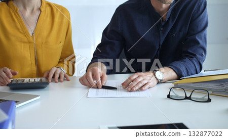 Business people engaging in a discussion about a contract while calculating costs during an office meeting, surrounded by documents, laptops, and financial paperwork. Audit and taxes theme in business Business people engaging in a discussion about a contract while calculating costs during an office meeting, surrounded by documents, laptops, and financial paperwork. Audit and taxes theme in business 132877022