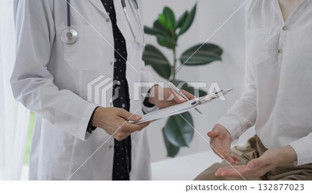 Doctor and a patient discussing something. The female physician, wearing a white medical coat is keeping clipboard with a medical notes besides a young woman during a consultation in the clinic Doctor and a patient discussing something. The female physician, wearing a white medical coat is keeping clipboard with a medical notes besides a young woman during a consultation in the clinic 132877023