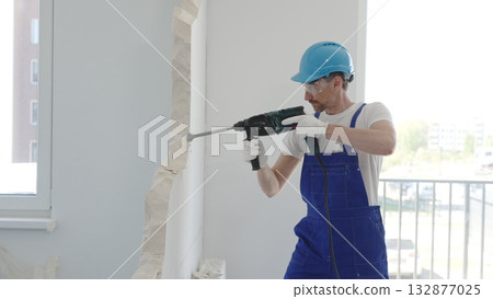 Male construction worker demolishing concrete wall with rotary hammer, wearing work overalls and blue hardhat is generating dust and debris during home renovation work, portrait horizontal view Male construction worker demolishing concrete wall with rotary hammer, wearing work overalls and blue hardhat is generating dust and debris during home renovation work, portrait horizontal view 132877025