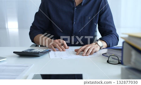 Businessman in a blue shirt examining financial documents at an office desk, using a calculator amidst paperwork and folders. Audit and taxes, business concept 132877030