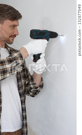 Middle aged male worker wearing checked shirt and white gloves, is drilling hole in a wall with cordless drill during home renovation work. Vertical portrait view 132877049