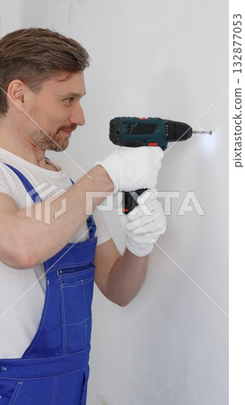 Man construction worker in a blue jumpsuit, safety goggles, and work gloves is screwing a screw into the wall using a cordless screwdriver, carrying out a careful home renovation. Vertical portrait Man construction worker in a blue jumpsuit, safety goggles, and work gloves is screwing a screw into the wall using a cordless screwdriver, carrying out a careful home renovation. Vertical portrait 132877053