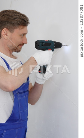 Man construction worker in a blue jumpsuit, safety goggles, and work gloves is screwing a screw into the wall using a cordless screwdriver, carrying out a careful home renovation. Vertical portrait Man construction worker in a blue jumpsuit, safety goggles, and work gloves is screwing a screw into the wall using a cordless screwdriver, carrying out a careful home renovation. Vertical portrait 132877081