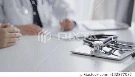 Black colored stethoscope over a tablet computer is in focus while doctor and a patient gesturing with their hands during a consultation in the clinic. Medicine concept Black colored stethoscope over a tablet computer is in focus while doctor and a patient gesturing with their hands during a consultation in the clinic. Medicine concept 132877082