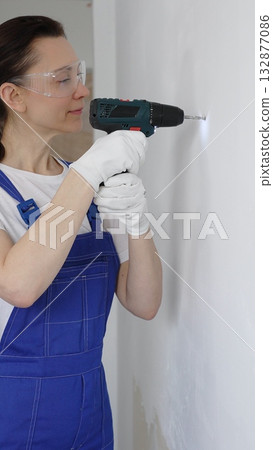 Professional female worker wearing blue overall, safety glasses and gloves, is using cordless drill to make hole in white wall during renovation work. Portrait view Professional female worker wearing blue overall, safety glasses and gloves, is using cordless drill to make hole in white wall during renovation work. Portrait view 132877086