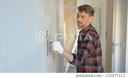 Male construction worker wearing gloves is carefully applying plaster to a wall using a hand tool, contributing to a home renovation or construction project 132877112