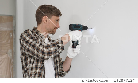 Man construction worker in checked shirt and safety gloves is screwing a screw into the wall using a cordless screwdriver, carrying out a careful home renovation. Horizontal portrait view Man construction worker in checked shirt and safety gloves is screwing a screw into the wall using a cordless screwdriver, carrying out a careful home renovation. Horizontal portrait view 132877124