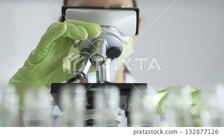 Close up of scientific precision involving green gloved hands adjusting microscope lens, blurred laboratory glassware highlighting medical investigation. Medicine, laboratory and science concept Close up of scientific precision involving green gloved hands adjusting microscope lens, blurred laboratory glassware highlighting medical investigation. Medicine, laboratory and science concept 132877126