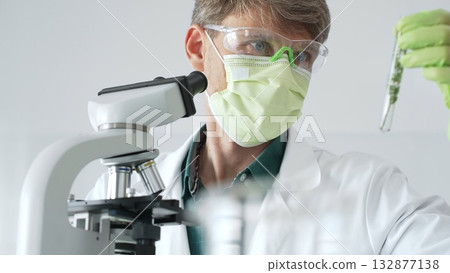 Adult male scientist wearing green mask, gloves and goggles using microscope and analyzing sample in test tube in a laboratory, developing new drugs and vaccines. Medicine and science concept Adult male scientist wearing green mask, gloves and goggles using microscope and analyzing sample in test tube in a laboratory, developing new drugs and vaccines. Medicine and science concept 132877138
