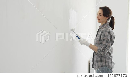 Woman wearing white gloves painting white wall with paint roller, experiencing home renovation and transforming living space 132877170