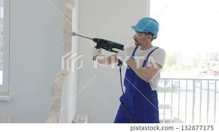 Construction worker demolishing concrete wall with rotary hammer, wearing protective safety gear, generating dust and debris during home renovation work, portrait horizontal view 132877175
