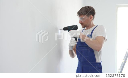 Middle aged male skilled worker drilling hole in white wall, wearing protective gear during home improvement project with cordless power tool Middle aged male skilled worker drilling hole in white wall, wearing protective gear during home improvement project with cordless power tool 132877178