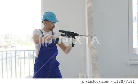 Male construction worker demolishing concrete wall with rotary hammer, wearing work overalls and blue hardhat is generating dust and debris during home renovation work, portrait horizontal view 132877181
