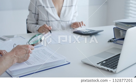 Two professional accountants working together, using a magnifying glass and a calculators, to audit financial reports in their brightly lit office. Audit and taxes in business 132877201