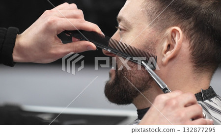 Skilled barber carefully trimming client's beard with precision scissors and sharp comb, working inside contemporary grooming salon 132877295