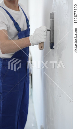 Professional construction worker wearing blue work overall is using a finishing trowel to carefully apply plaster on a wall, demonstrating expertise in home renovation and interior finishing Professional construction worker wearing blue work overall is using a finishing trowel to carefully apply plaster on a wall, demonstrating expertise in home renovation and interior finishing 132877300