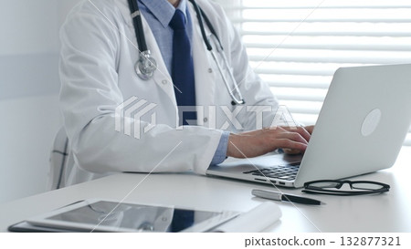 Medical doctor wearing lab coat and stethoscope is sitting at desk and typing on laptop, with eyeglasses and pen nearby, in medical office. Medicine and health care concept 132877321