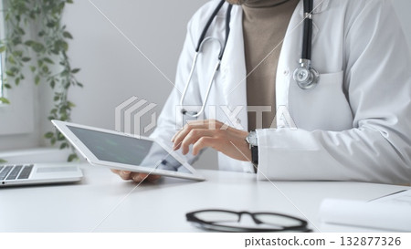 Doctor man wearing lab coat and stethoscope using digital tablet in modern medical office, browsing patient information or conducting online consultation. Medicine and health care 132877326