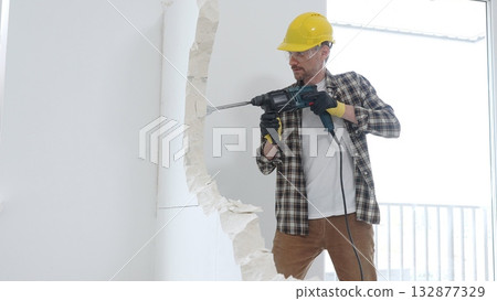 Male construction worker wearing beige checkered shirt, yellow hard hat and protective gloves, is demolishing white wall with rotary hammer drill, generating dust, medium long shot. Renovation project Male construction worker wearing beige checkered shirt, yellow hard hat and protective gloves, is demolishing white wall with rotary hammer drill, generating dust, medium long shot. Renovation project 132877329
