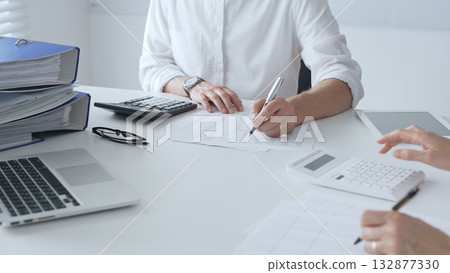 Two unrecognizable accountants collaborating in a well lit office, analyzing financial data and preparing reports with calculators and pens. Audit and taxes concept Two unrecognizable accountants collaborating in a well lit office, analyzing financial data and preparing reports with calculators and pens. Audit and taxes concept 132877330