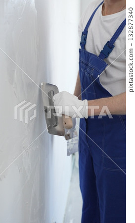 Professional construction worker wearing blue work overall is using a finishing trowel to carefully apply plaster on a wall, demonstrating expertise in home renovation 132877340