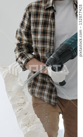 Close up of unknown male construction worker wearing beige checkered shirt and protective gloves, is demolishing white wall with rotary hammer drill, generating dust. Renovation project 132877428