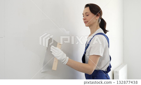 Woman construction worker wearing protective gloves and blue construction coveralls, spreading plaster smoothly across wall using professional drywall taping knife during renovation project Woman construction worker wearing protective gloves and blue construction coveralls, spreading plaster smoothly across wall using professional drywall taping knife during renovation project 132877438