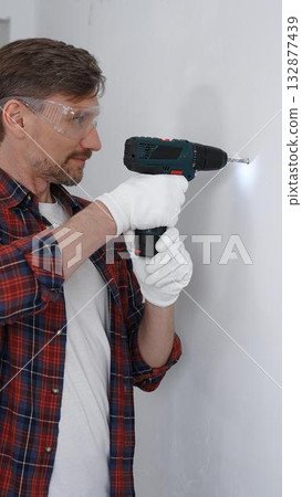 Middle aged male professional worker wearing red checked shirt and white gloves, is drilling hole in a wall with cordless drill during home renovation work. Vertical portrait view Middle aged male professional worker wearing red checked shirt and white gloves, is drilling hole in a wall with cordless drill during home renovation work. Vertical portrait view 132877439