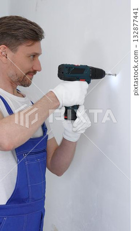 Professional male construction worker in a blue jumpsuit, safety goggles, and work gloves is screwing a screw into the wall using a cordless screwdriver, carrying out a careful home renovation 132877441