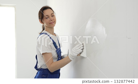 Construction worker woman spreading finishing putty on white wall, wearing protective gloves while smoothing surface with putty knife for precise home repair 132877442