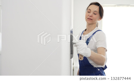 Professional female construction worker wearing blue coveralls is using a finishing trowel to carefully apply plaster on a wall, demonstrating expertise in home renovation 132877444
