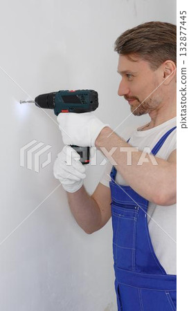 Man construction worker in a blue jumpsuit, safety goggles, and work gloves is screwing a screw into the wall using a cordless screwdriver, carrying out a careful home renovation. Vertical portrait 132877445
