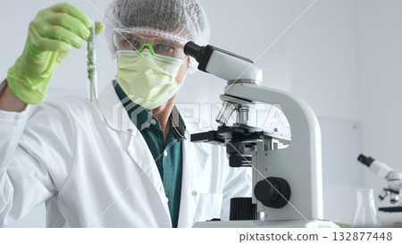 Adult male scientist wearing green protective gear looking at plant samples in test tubes and using a microscope in modern laboratory for botanical research. Medicine, healthcare and science concept Adult male scientist wearing green protective gear looking at plant samples in test tubes and using a microscope in modern laboratory for botanical research. Medicine, healthcare and science concept 132877448