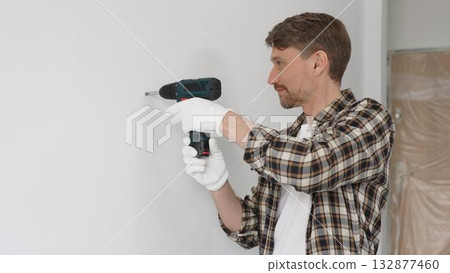 Man construction worker in checked shirt and safety gloves is screwing a screw into the wall using a cordless screwdriver, carrying out a careful home renovation. Horizontal portrait view 132877460
