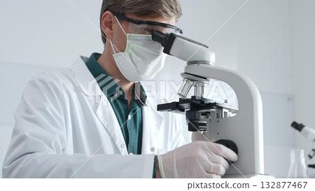 Adult male scientist wearing white mask, lab coat and protective glasses using microscope, doing scientific research in modern laboratory. Medicine, health care and science concept Adult male scientist wearing white mask, lab coat and protective glasses using microscope, doing scientific research in modern laboratory. Medicine, health care and science concept 132877467