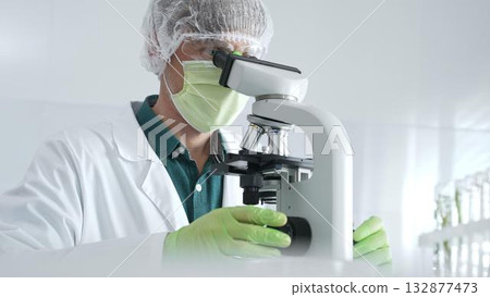 Medical scientist wearing protective mask, cap and green gloves using microscope doing scientific research in laboratory, developing vaccine or medication. Medicine, healthcare and science concept Medical scientist wearing protective mask, cap and green gloves using microscope doing scientific research in laboratory, developing vaccine or medication. Medicine, healthcare and science concept 132877473