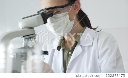 Female scientist examining microscope slide, wearing protective gear while conducting medical research in sterile laboratory environment. Medicine, healthcare and science concept Female scientist examining microscope slide, wearing protective gear while conducting medical research in sterile laboratory environment. Medicine, healthcare and science concept 132877475