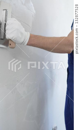 Professional construction worker wearing blue work overall is using a finishing trowel to carefully apply plaster on a wall, demonstrating expertise in home renovation and interior finishing Professional construction worker wearing blue work overall is using a finishing trowel to carefully apply plaster on a wall, demonstrating expertise in home renovation and interior finishing 132877528