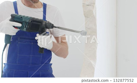 Unrecognizable male construction worker wearing blue overalls and protective white gloves, is demolishing white wall with rotary hammer drill, generating dust, close up horizontal view. Renovation Unrecognizable male construction worker wearing blue overalls and protective white gloves, is demolishing white wall with rotary hammer drill, generating dust, close up horizontal view. Renovation 132877552