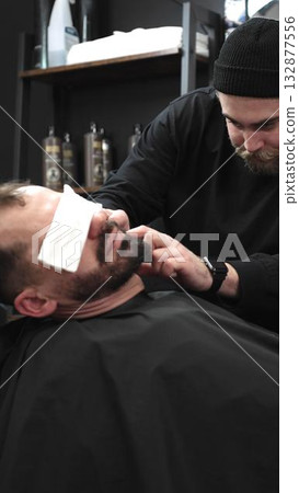Professional barber carefully shaving client's beard with straight razor, applying gel and using hot towel in barbershop setting, vertical view 132877556