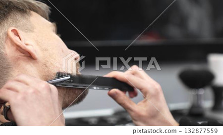 Professional barber grooming client's beard with precision, using comb and razor within stylish contemporary barbershop setting Professional barber grooming client's beard with precision, using comb and razor within stylish contemporary barbershop setting 132877559