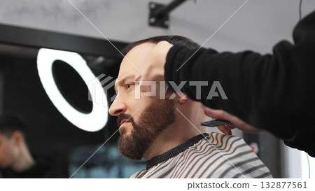 Skilled barber carefully grooming client's beard with electric razor, creating precise, trendy look inside contemporary barbershop setting 132877561