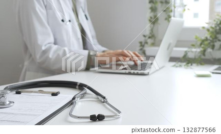 Stethoscope and medical records are on the desk opposite the doctor is typing on a laptop with a in a medical office, suggesting online healthcare or telemedicine services. Medicine concept 132877566