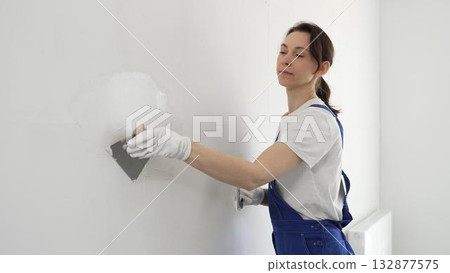Construction worker woman spreading finishing putty on white wall, wearing protective gloves while smoothing surface with putty knife for precise home repair Construction worker woman spreading finishing putty on white wall, wearing protective gloves while smoothing surface with putty knife for precise home repair 132877575